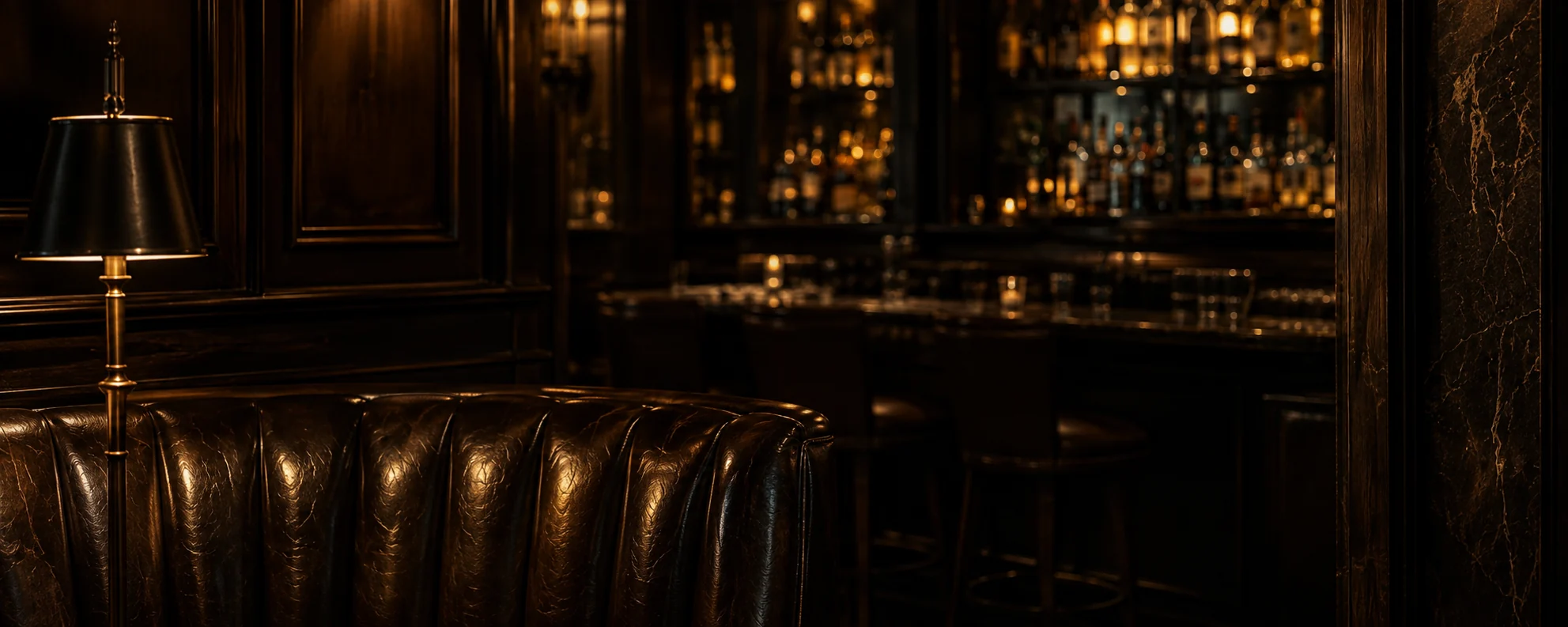 Dark bar lounge after closing — tufted leather banquette and brass lamp facing a lit back bar