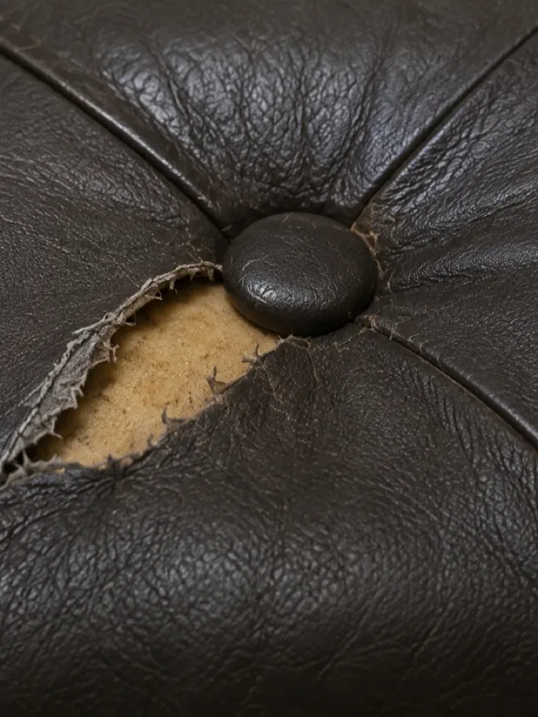 An open tear beside a button on dark buttoned leather upholstery with foam visible inside