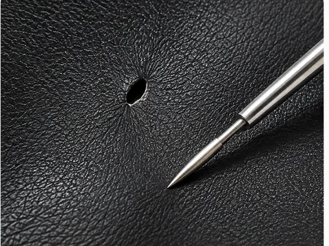 A small puncture in a black vinyl panel with a fine repair tool angled into it, the surrounding grain intact and undamaged