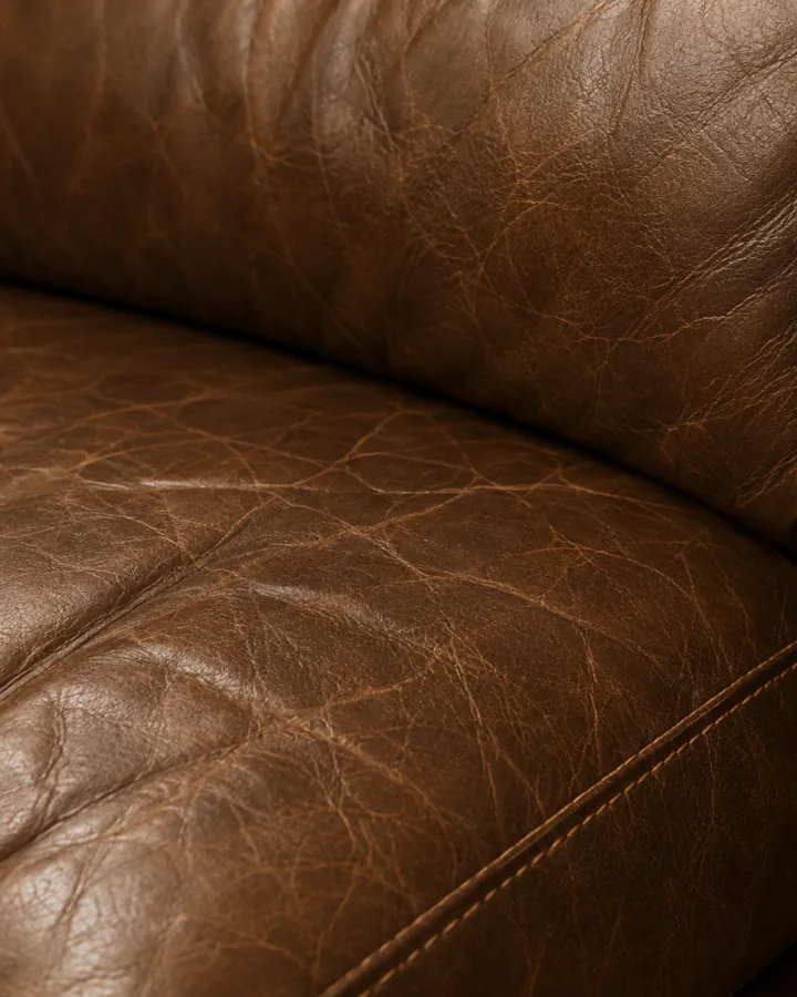 A worn brown full grain leather sofa arm and seat, creased and marked, the growth lines of the hide still showing.