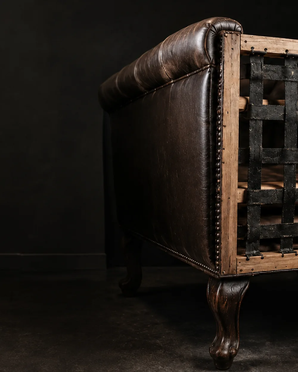 The end of a buttoned brown leather sofa with the seat open, black webbing woven in a lattice across the timber frame above a carved cabriole leg