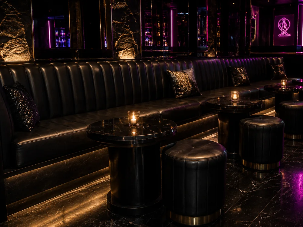 Long channel-tufted black leather wall bench with round stools in a nightclub