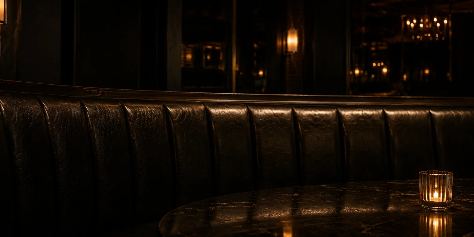 Long channel-tufted black leather banquette and marble table with a lit candle in a dark nightclub lounge