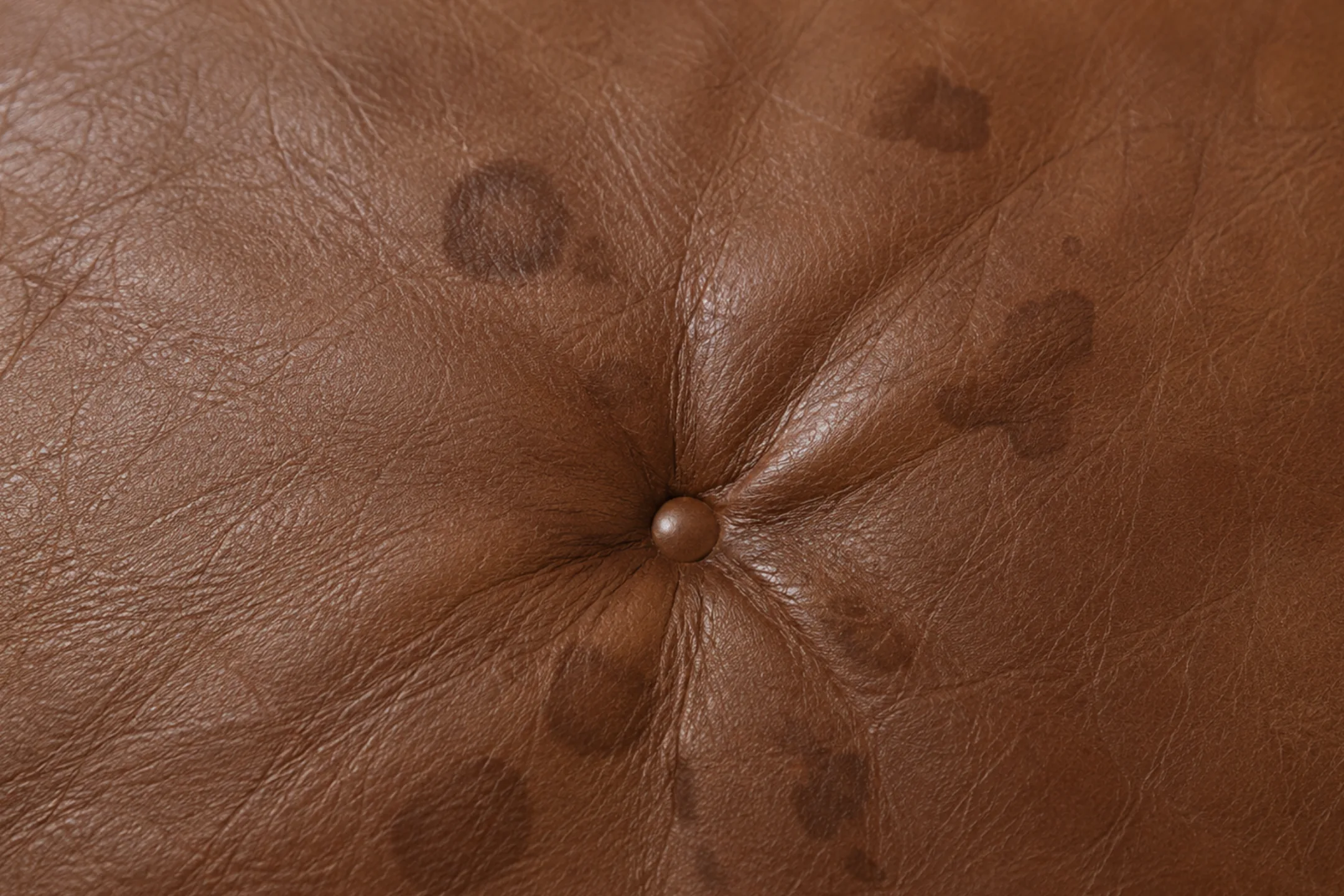 Dark stain blotches spread around a button tuft on brown semi-aniline leather, absorbed into the dye beneath the finish
