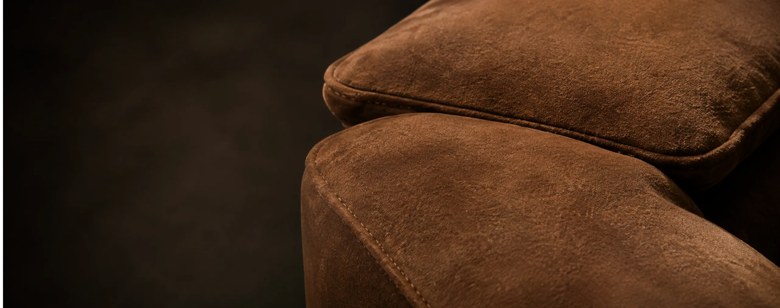 The arm and seat cushion of a tan suede sofa lit from one side against a dark room, the nap reading light on the roll and dark in the fold