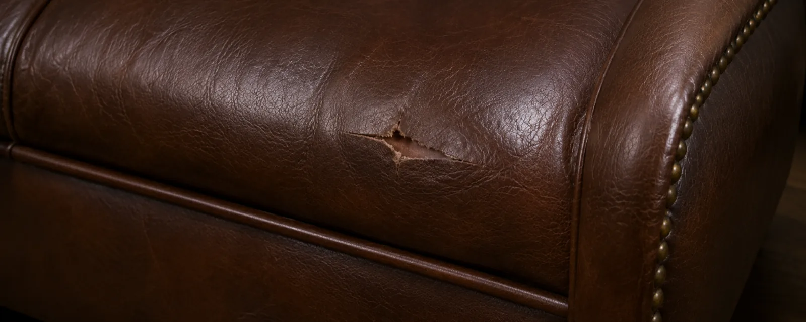 A torn hole in the rolled arm of a dark brown studded top-grain leather sofa