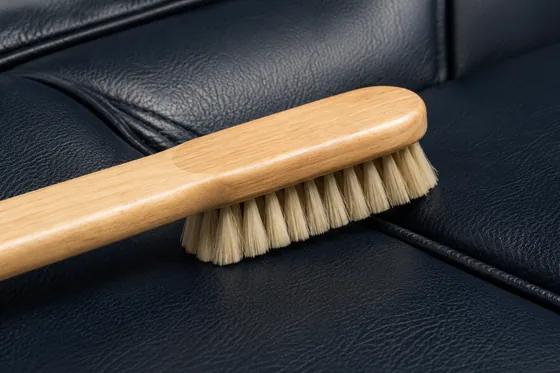 A wooden-handled soft bristle brush being drawn along the stitched seam of a dark vinyl seat