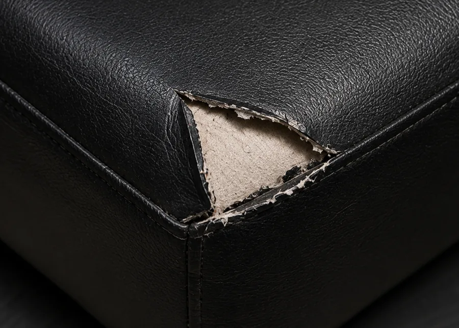 The corner of a black vinyl cushion torn open in a triangle, the pale woven backing clearly visible inside while the vinyl all around the tear is smooth and undamaged