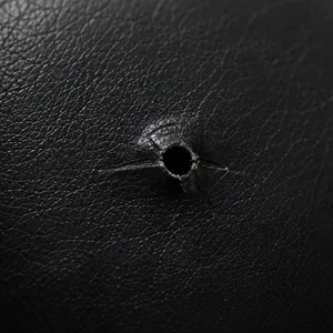 A small round puncture hole punched through black vinyl upholstery, the film around it still smooth