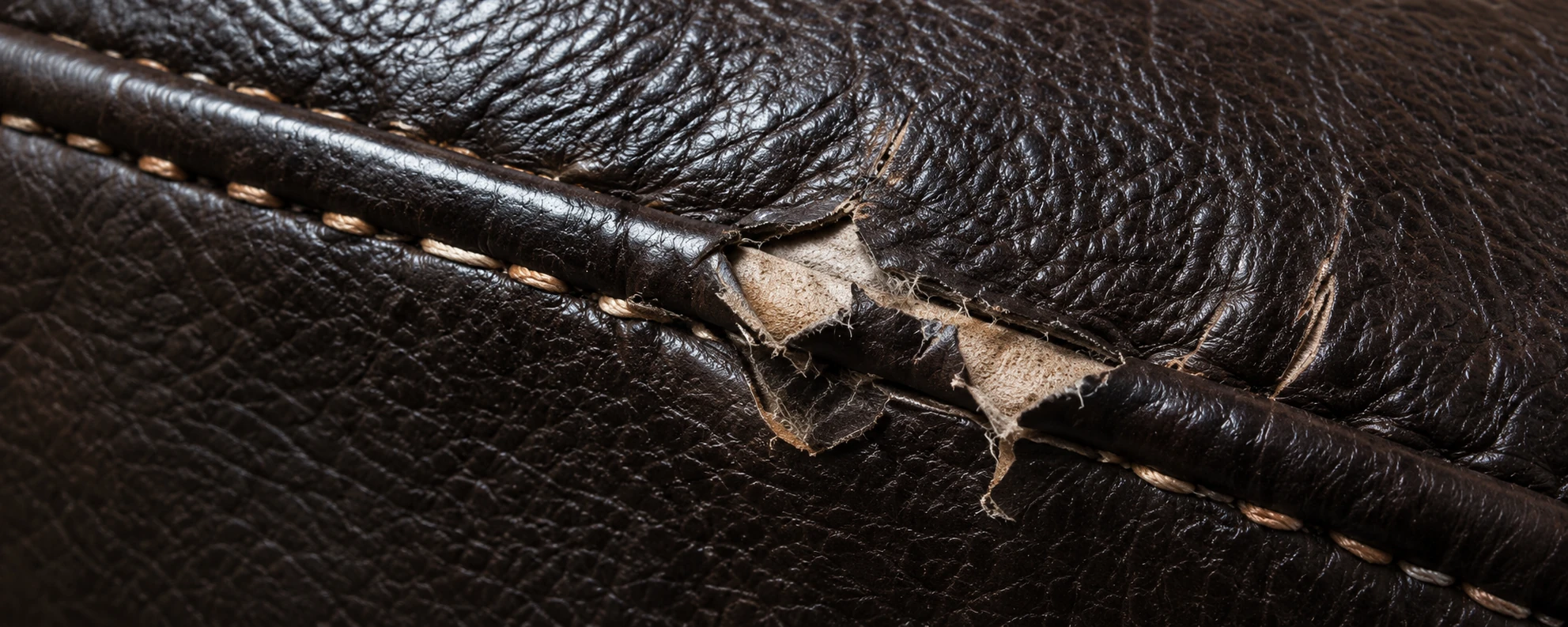 Brown leather torn open beside a piped seam, exposing the pale backing underneath