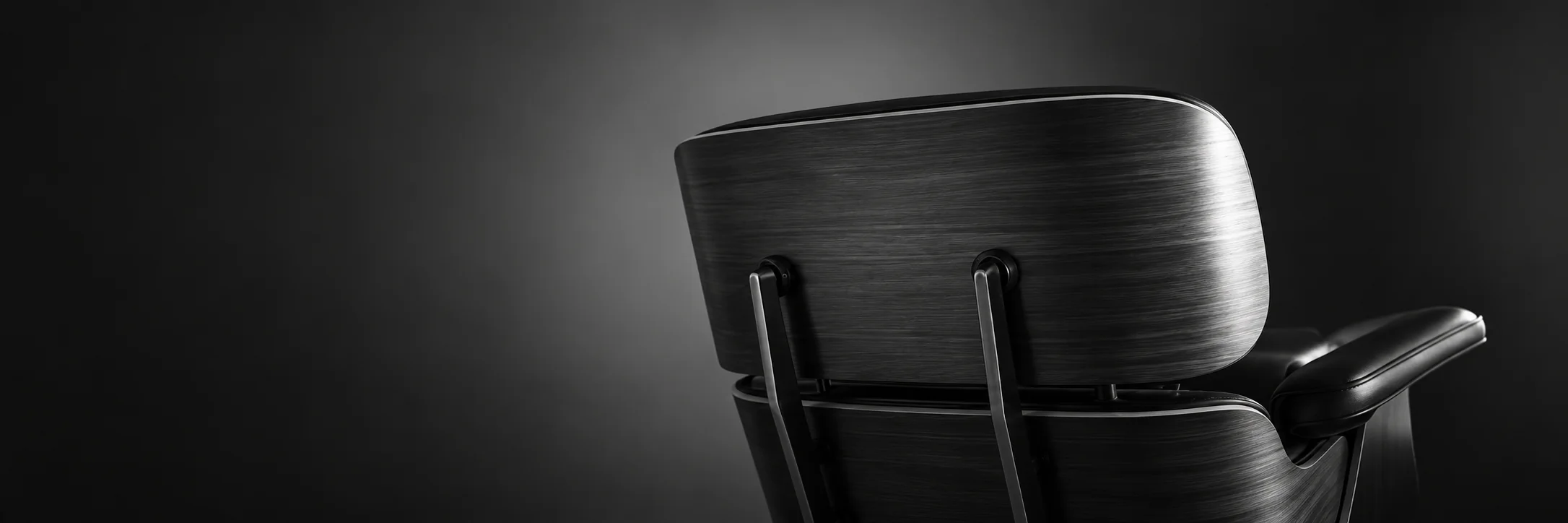 The walnut shell and black leather armrest of an Eames lounge chair lit against a dark background