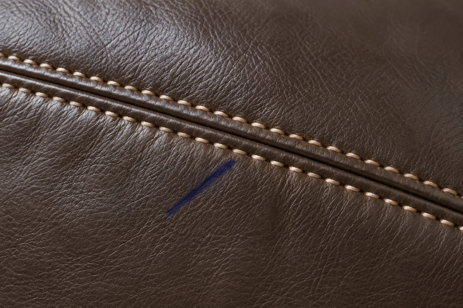 A short blue ink mark on dark brown leather beside a contrast-stitched seam