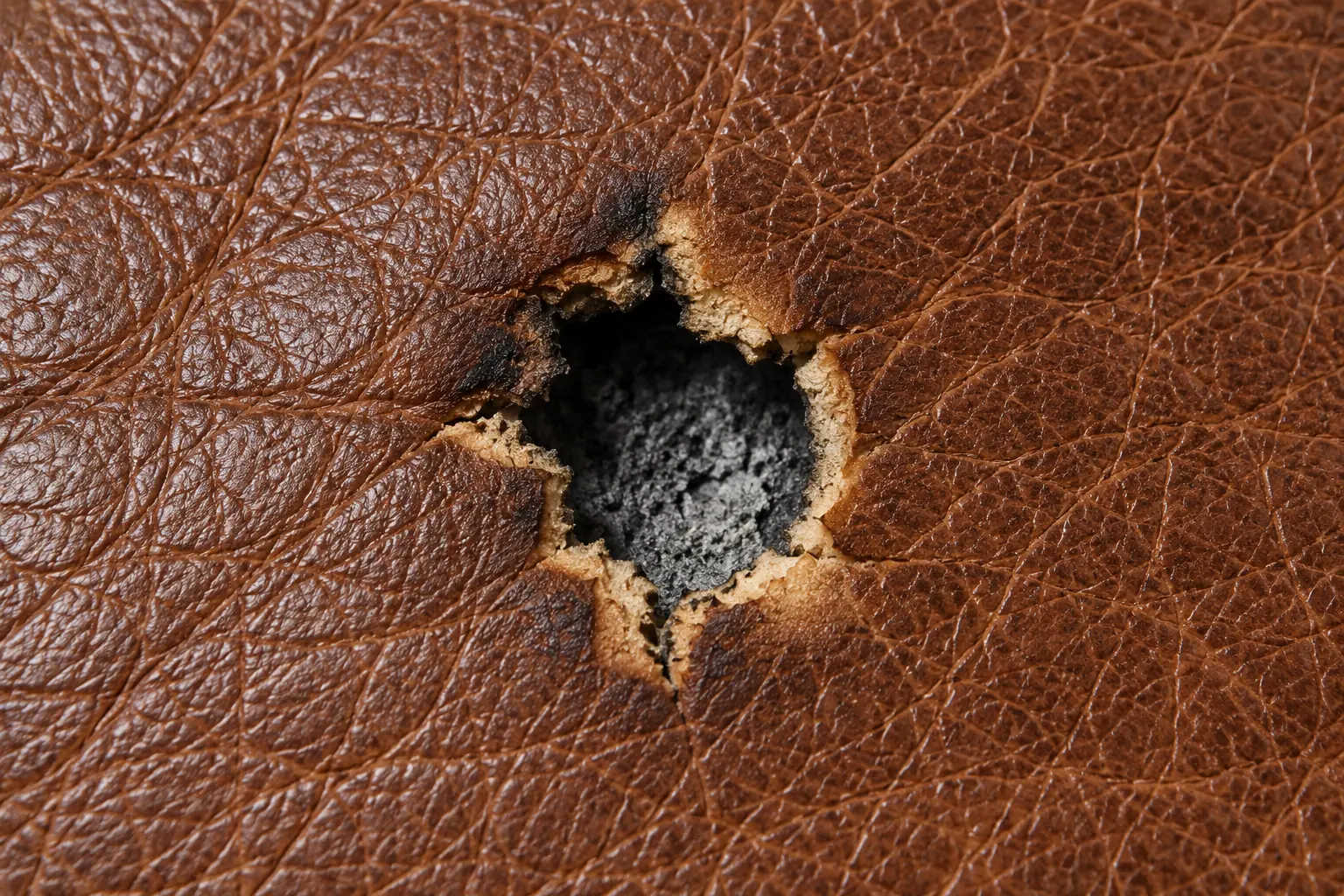 Burn hole through brown leather with a charred rim and dark foam showing underneath