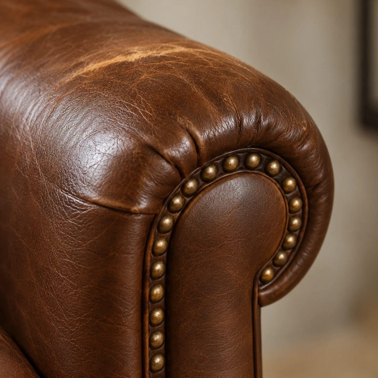 A close view of a brown leather chair arm where the piped seam runs beside a line of antique brass nailheads