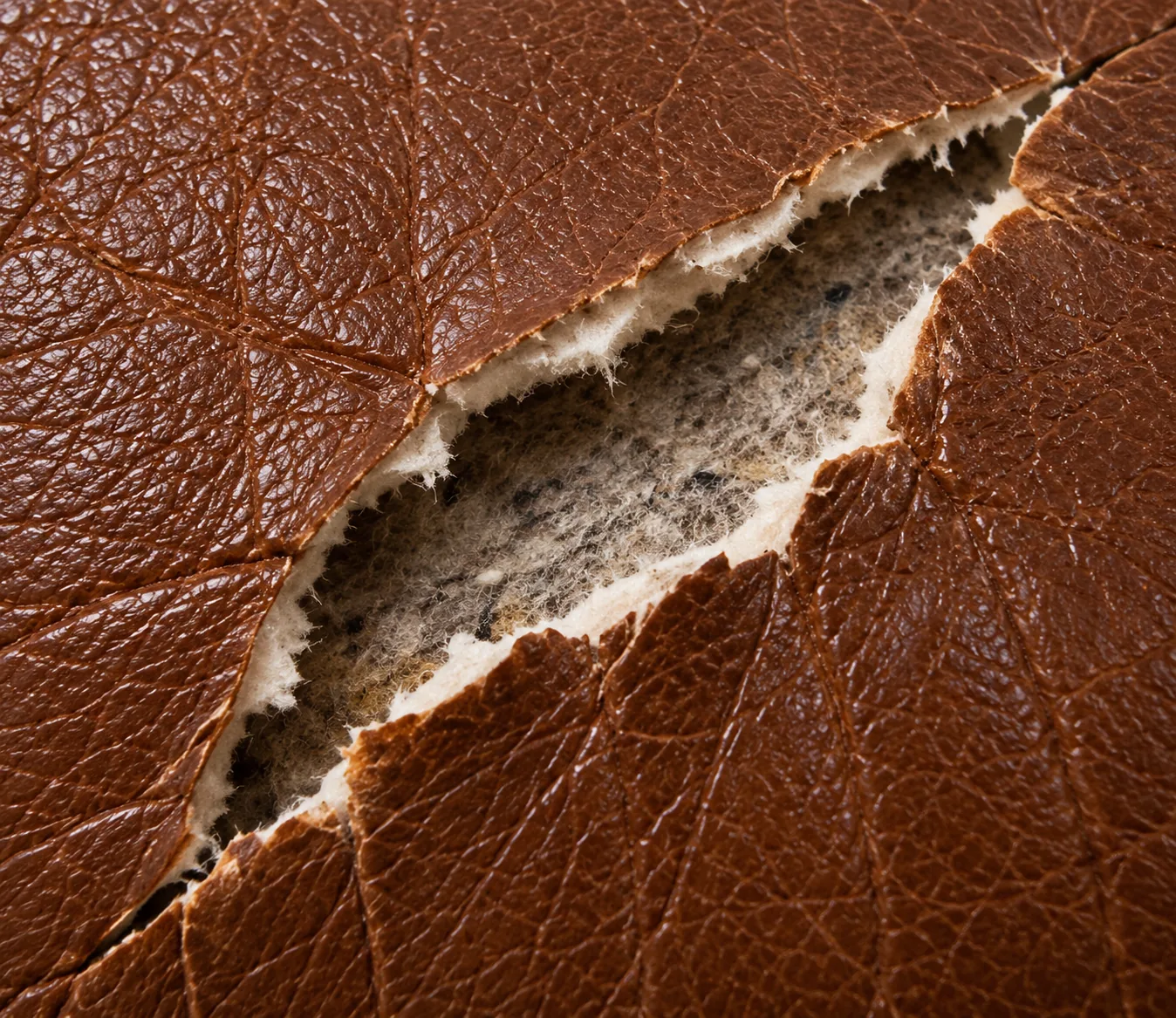 A rip in brown leather: a wide opening with the edges pulled apart and pale backing showing through