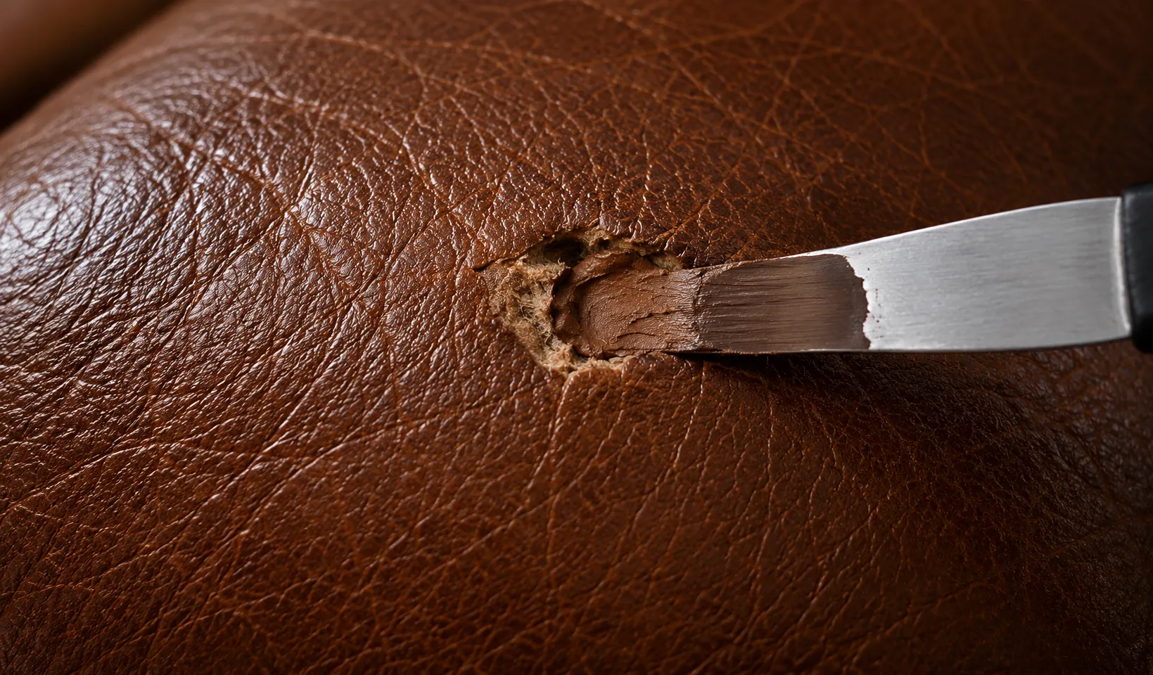 Flexible filler being drawn level across a backed rip in brown leather with a spatula