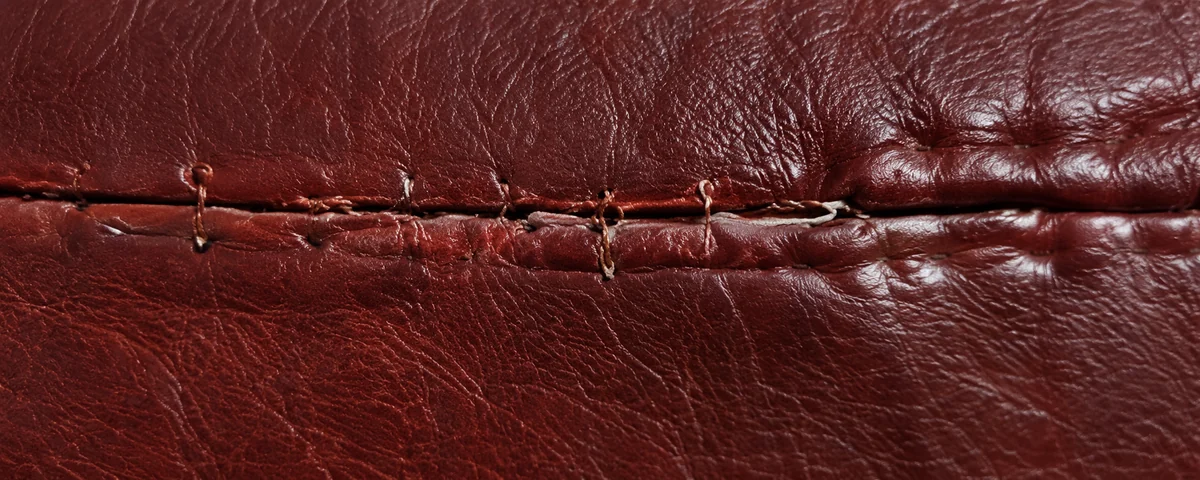 A burgundy leather seam closed with coarse hand stitching at uneven spacing, already lifting away