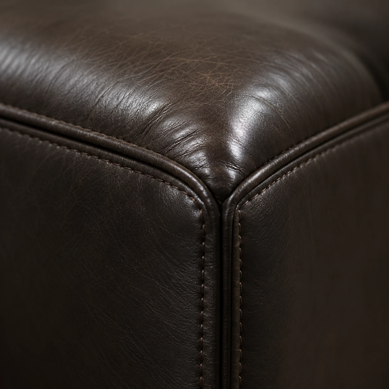 Close-up of a dark brown leather sofa corner where two welted seams meet on a square arm