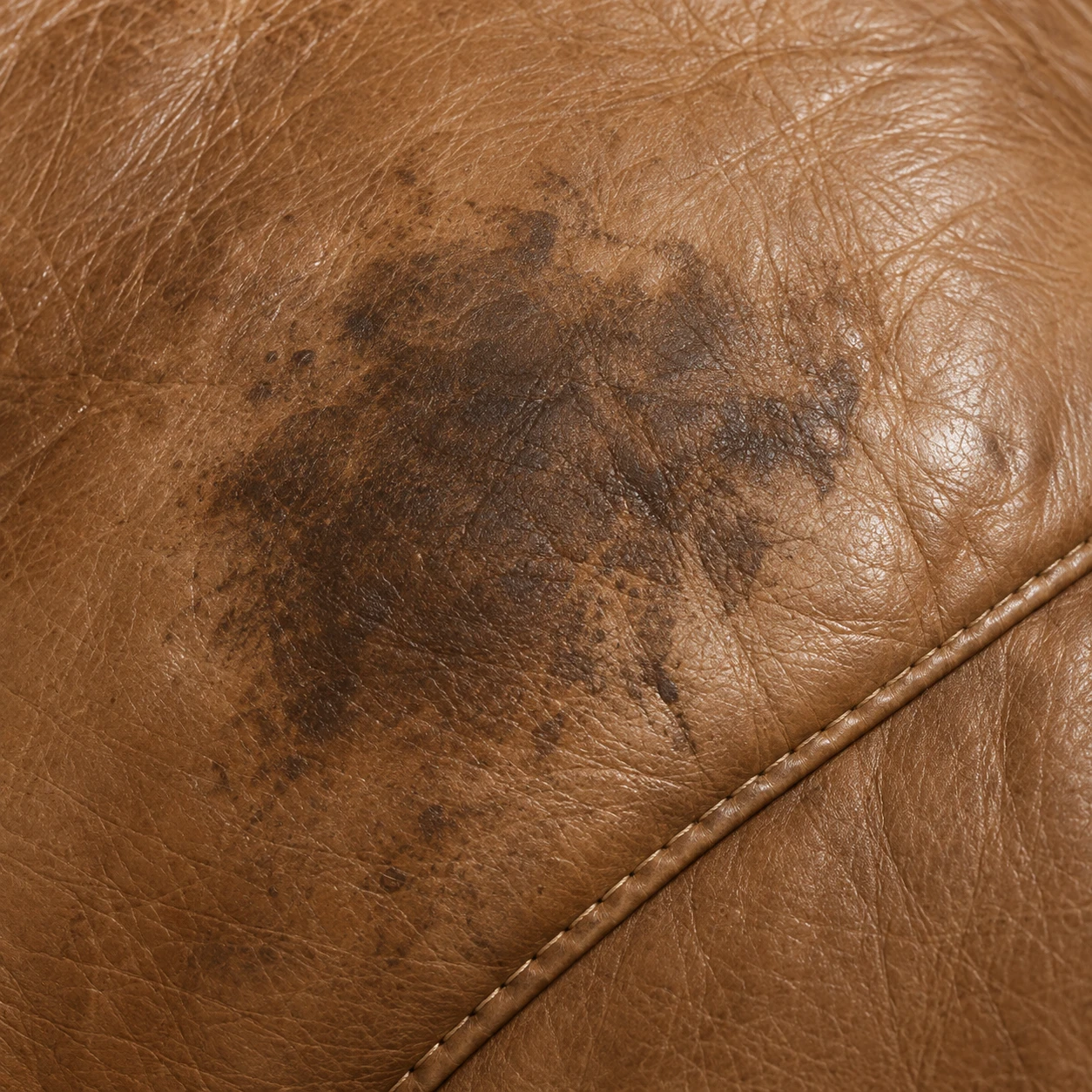 A dark set-in stain on a tan leather seat beside a stitched seam