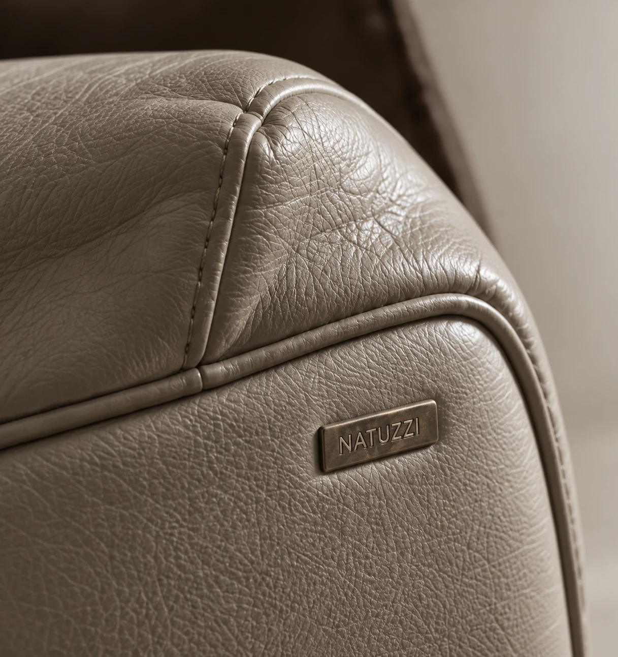 Close-up of taupe Natuzzi leather showing the piped seam, topstitching and the brass Natuzzi badge on the arm