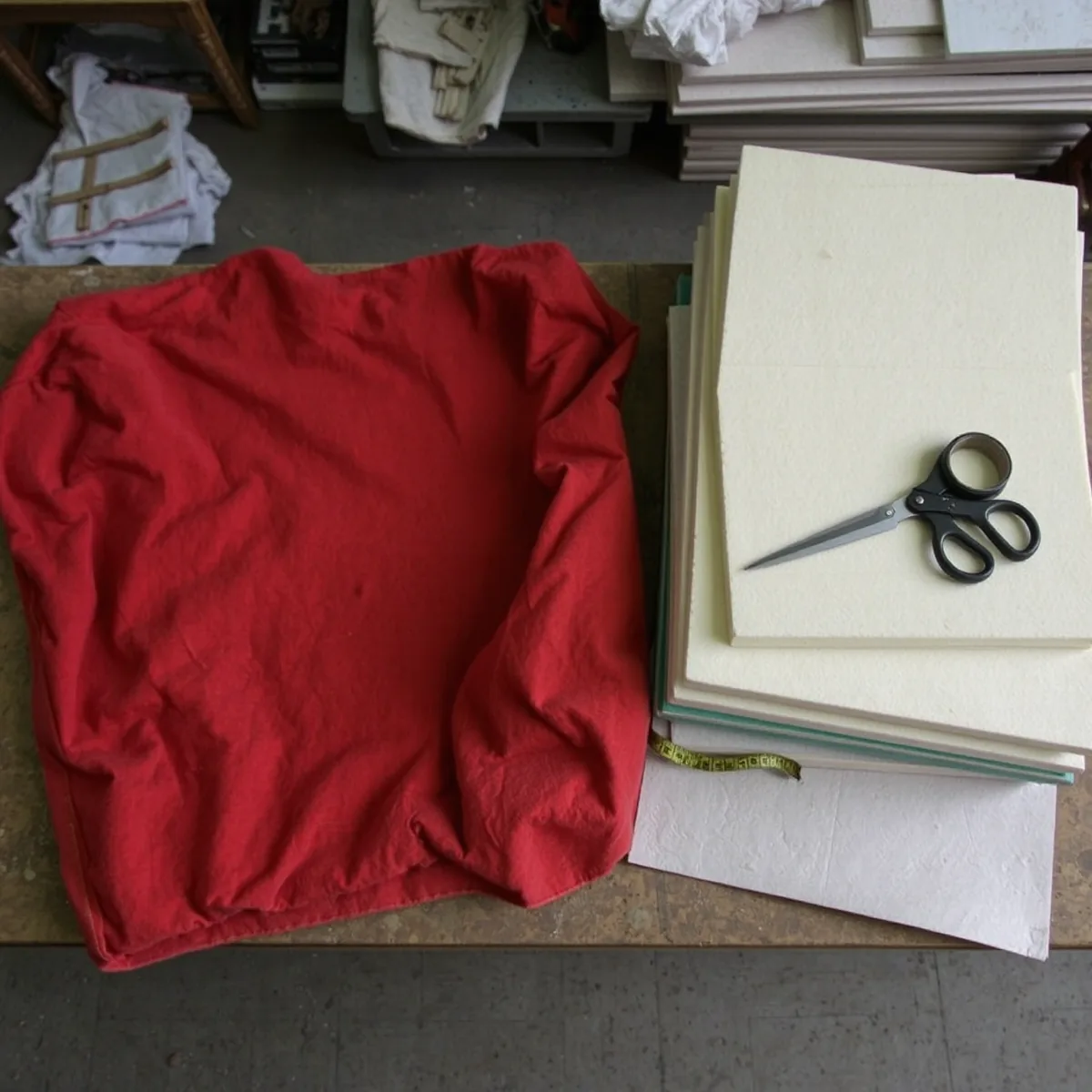 An old faded red theatre seat cover panel laid flat on a workbench beside a stack of foam and wadding layers, shears and a tape measure across them