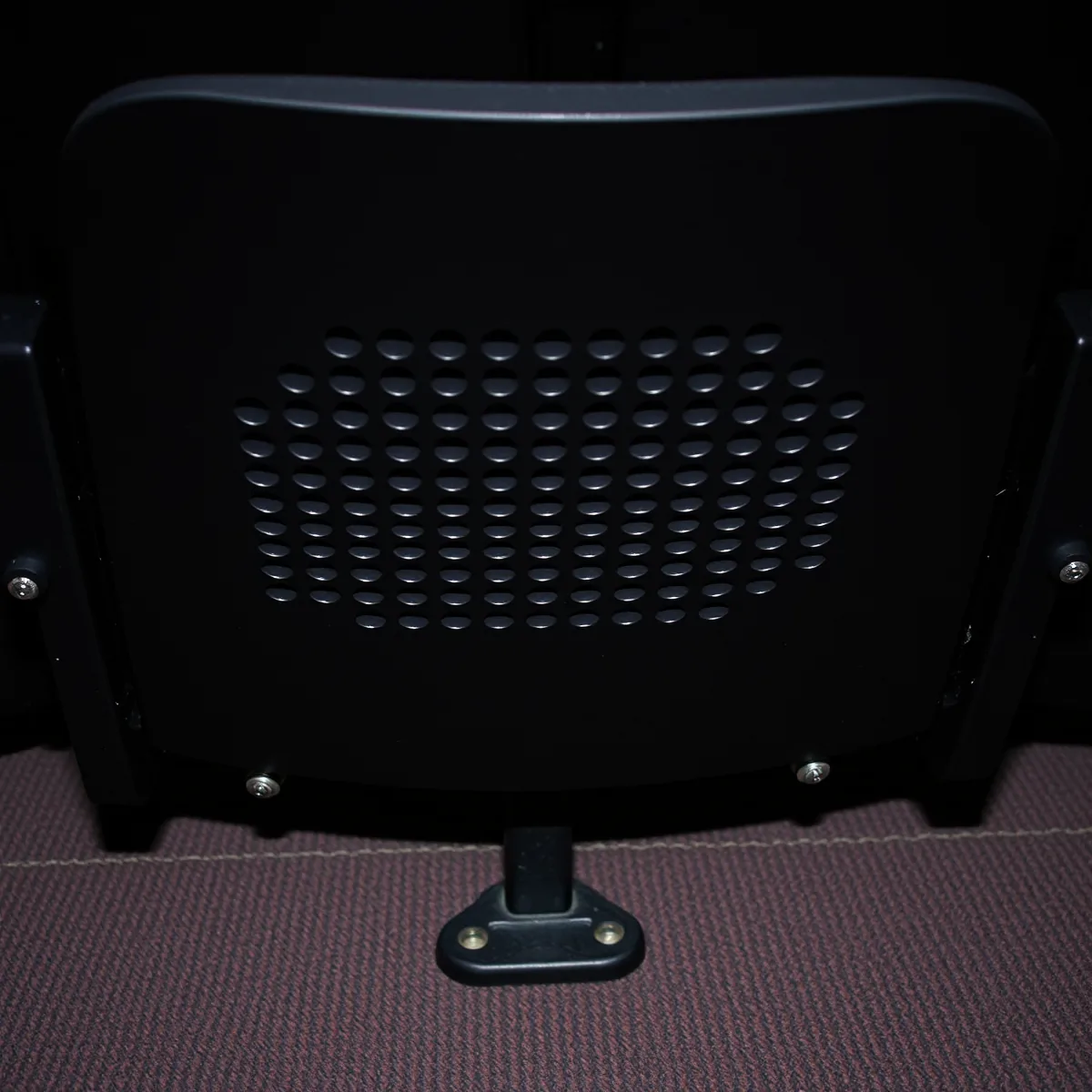 Underside of a folded-up theatre seat bottom, a dark panel pierced with a grid of small round ventilation holes above a cast iron standard